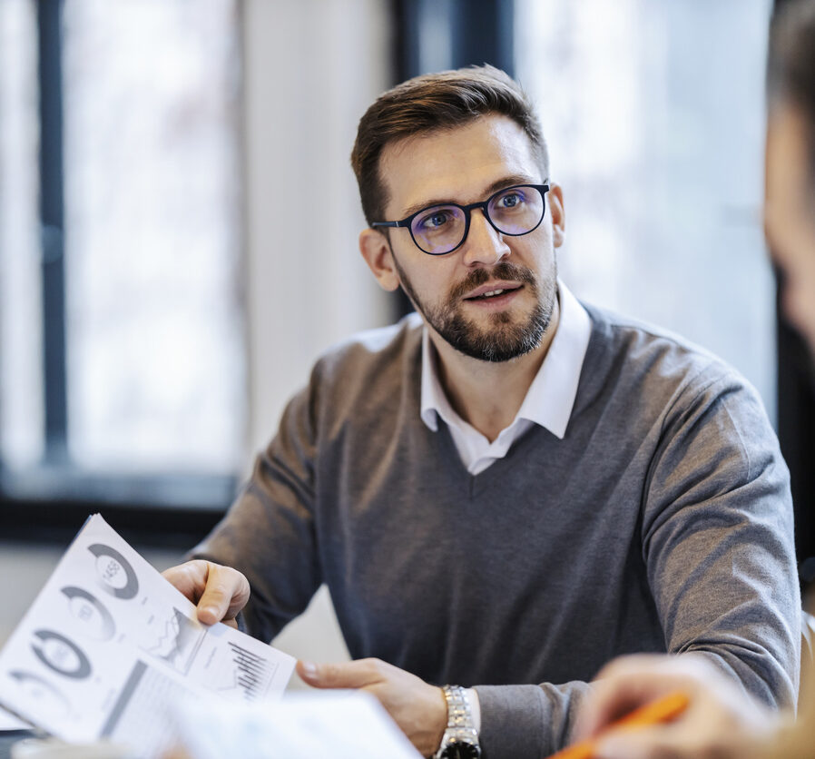 A manager is sitting with his colleague on a briefing at meeting room and discussing statistics and charts on paperwork.