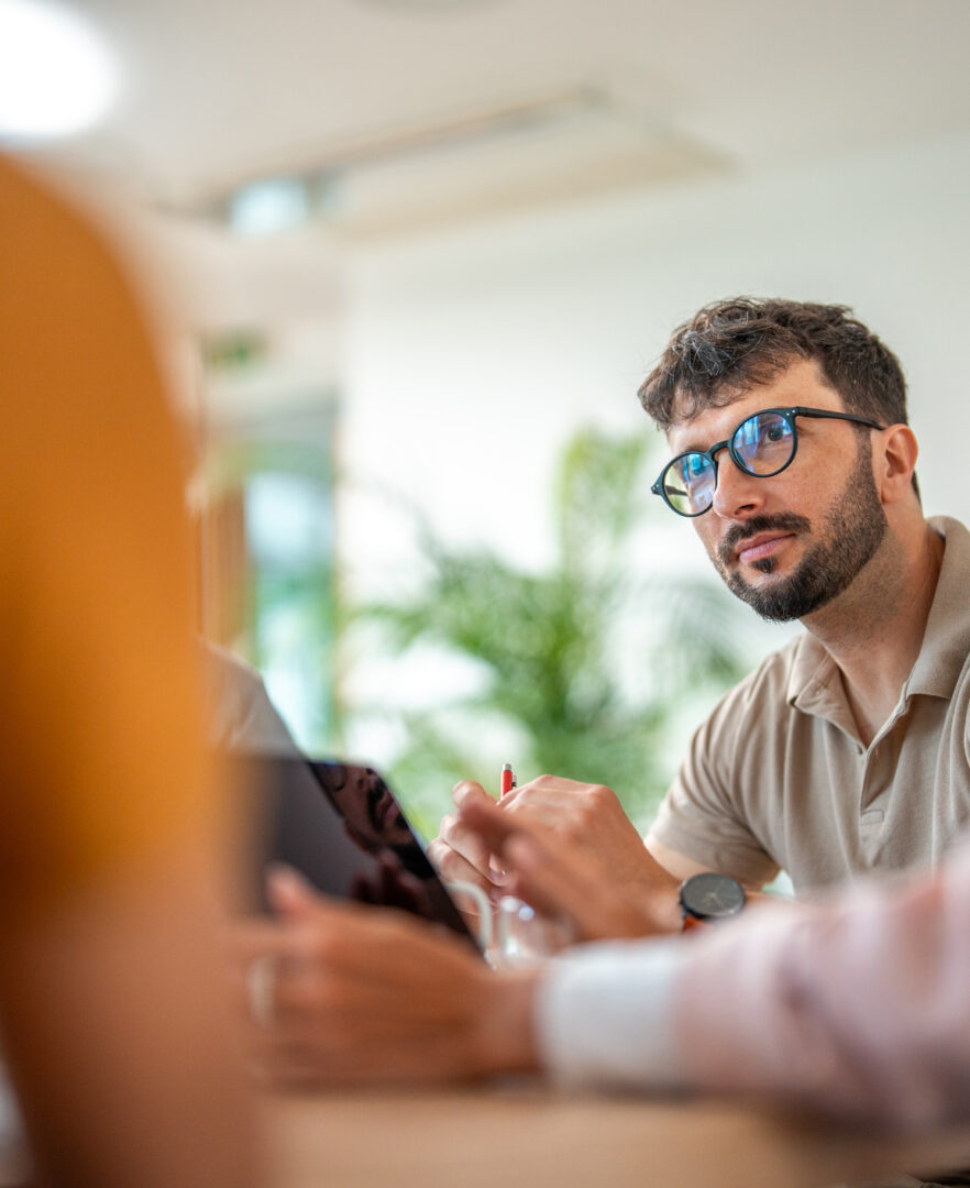 Caucasian male in casual business attire leads a meeting, actively engaging with colleagues of diverse backgrounds in a bright office setting.